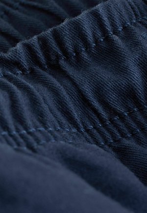 Teksturēta tumši zila auduma ar savāktām šuvēm, kas izceļ mīkstu un elastīgu materiālu. Uzsvars uz elastīgo jostasvietu.