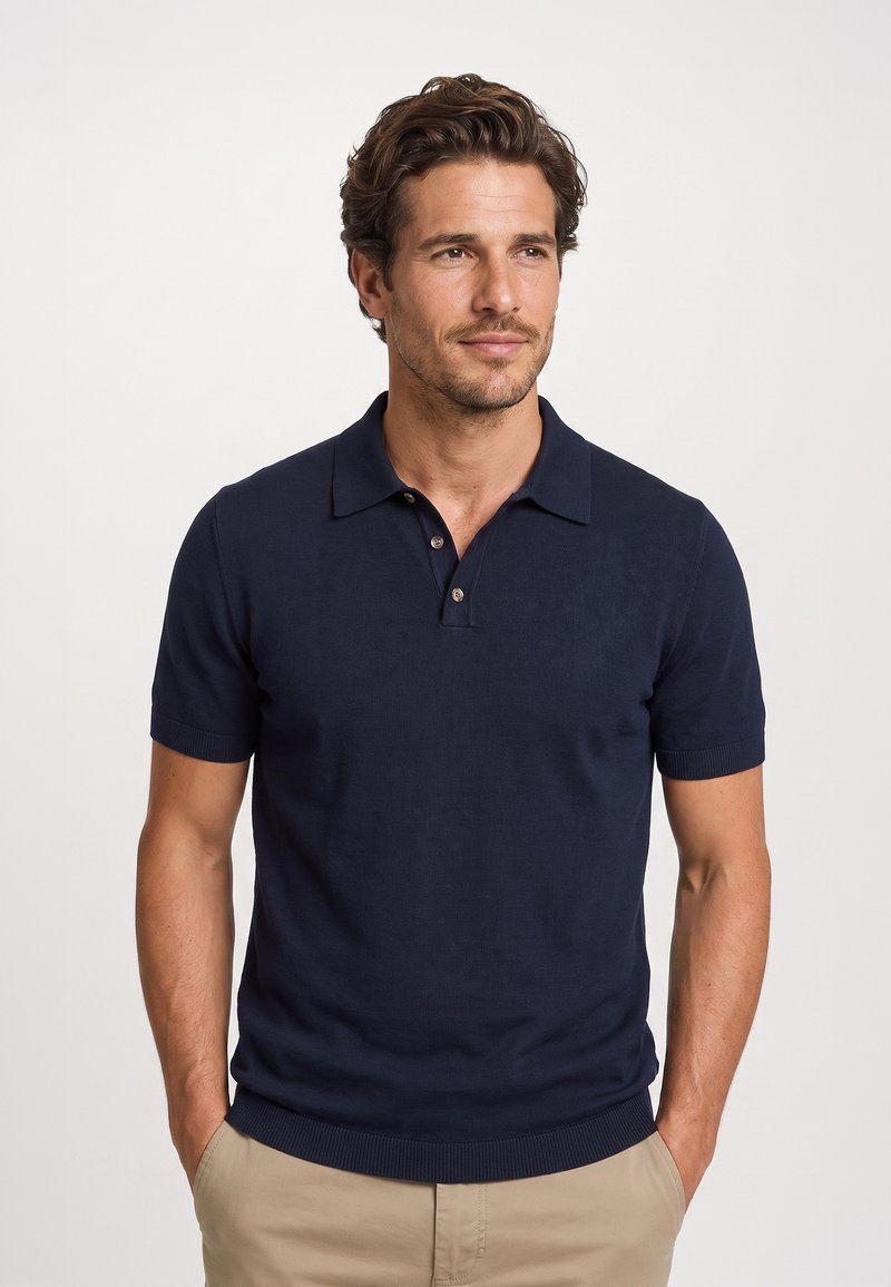 Mann mit kurzen lockigen Haaren, der ein dunkelblaues kurzärmeliges Poloshirt und beige Hosen trägt, steht mit den Händen in den Taschen vor einem schlichten Hintergrund.