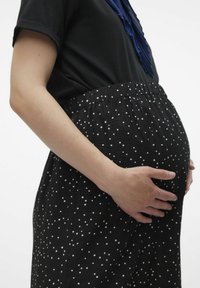 Robe de maternité à pois noirs avec une taille élastique, en tissu doux ; personne tenant un ventre de grossesse visible, vue de profil.