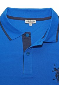 Blå pikétröja i texturerat tyg, med en mörkblå krage och två blå knappar. Svart broderad logotyp av en polospelare på vänster sida.