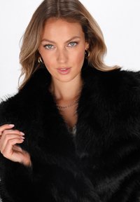 Kvinde med lysebrunt hår iført guldørehængere og en sort pelsjakke i fake fur, som hun holder kraven på med den ene hånd.
