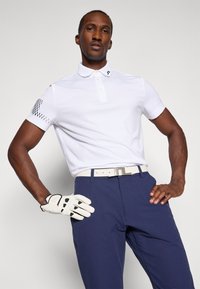 Hvit kortermet polo med knappestolpe og strukturert mønster på venstre erm, kombinert med marineblå bukser og en hvit golfhanske.