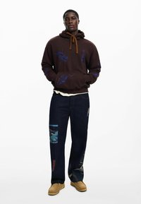 Brauner Hoodie mit blauen Grafiken und einer Fronttasche, kombiniert mit dunklen Jeans aus Denim, die verschiedene Stoffpatches aufweisen. Beige Schuhe runden das Outfit ab.