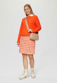 Strukturierter orangener Pullover, gemusterter Rock im pink-weißen Hahnentrittmuster, beigefarbene Schultertasche und cremefarbene Schnürstiefel. Neutraler Hintergrund.