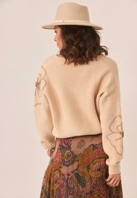 Suéter de punto beige con textura acanalada, adornado con bordados florales en las mangas, combinado con una falda colorida y estampada. La modelo lleva un sombrero beige.