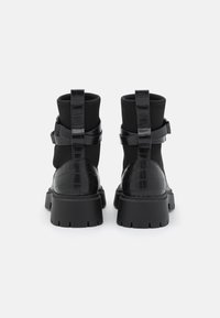 Bottes noires à chevilles avec une tige synthétique à texture embossée effet crocodile, des panneaux élastiques et une semelle en caoutchouc épaisse. Deux sangles à l'arrière.