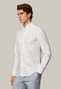 Mladić smeđe kose, obučen u bijelu košulju na kopčanje i svijetloplave prugaste hlače, stoji s jednom rukom u džepu i gleda lijevo.