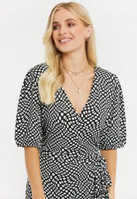 Vestido negro ajustado con un patrón de lunares blancos, mangas cortas con volumen y un escote en V pronunciado. Textura suave con un detalle de cintura con lazo.