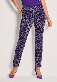 Vrouw in paarse leopardprint strakke broek, gecombineerd met paarse hoge hakken en een paarse top, staand tegen een effen achtergrond.