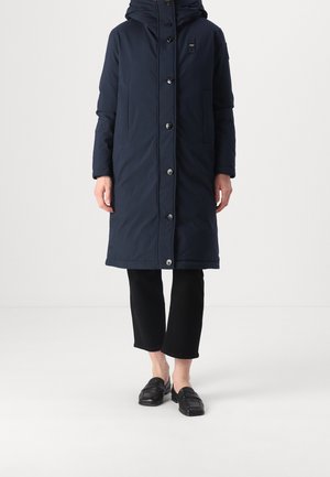 Person trägt einen langen marineblauen gesteppte Mantel mit Knöpfen, schwarze kurze Hosen und schwarze Loafer und steht vor einem weißen Hintergrund.