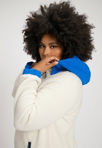 Chaqueta de forro polar en crema con capucha azul, con una superficie texturizada y puños en contraste; cierre con cremallera y sutiles detalles en negro.