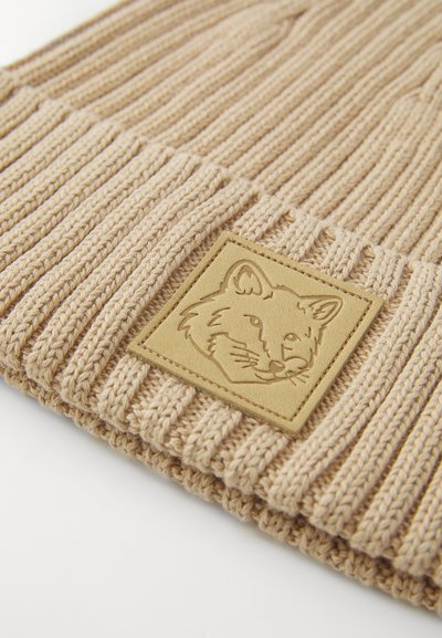 Bonnet en tricot beige avec texture côtelée et un patch en cuir carré, arborant un design de tête de renard. Matière douce et chaude.