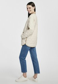 Beżowa pikowana kurtka z okrągłym dekoltem, bocznymi kieszeniami i kontrastowym jasnobrązowym wykończeniem, w połączeniu z niebieskimi dżinsami i białymi trampkami.