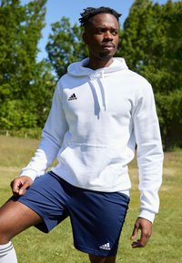 Hvit bomullshoodie med lomme foran og sort Adidas-logo, kombinert med marineblå treningsshorts med hvit logo og elastisk midjebånd.