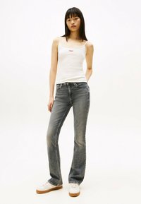 Top sin mangas blanco con letras rojas, combinado con jeans acampanados de mezclilla azul desgastada y zapatillas blancas con suelas beige. Diseño sencillo y textura suave.