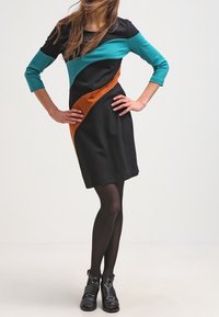 Robe noire avec des manches trois-quarts teal, un motif diagonal teal et orange, portée avec des bottines noires et des collants.