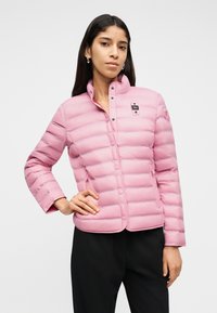 Γυναίκα με μακριά σκούρα μαλλιά που φοράει ανοιχτό ροζ ανάγλυφο μπουφάν με κουμπιά, σε συνδυασμό με μαύρο παντελόνι, όρθια με το ένα χέρι στη μέση.