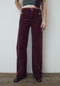 Bordowe szerokie spodnie z sztruksu z dwoma przednimi kieszeniami i złotymi guzikami. Wykonane z fakturowanego materiału z prostą linią nogawek, noszone z szarą bluzką.