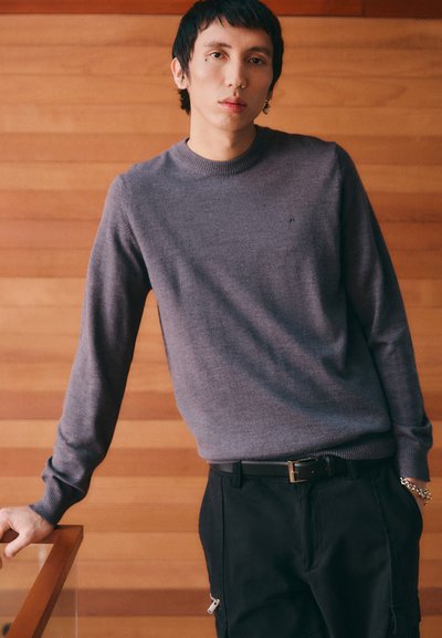 Pull en maille gris avec un col rond et des manches longues, associé à un pantalon noir avec des zips sur les côtés et une ceinture en cuir. Design simple.