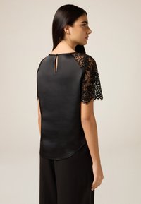 Czarna bluzka z satynowym korpusem i koronkowymi rękawami, z ozdobnym wycięciem na plecach i falistym wykończeniem rękawów. Gładka faktura, dopasowany krój.