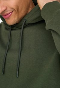 Mørkegrøn sweat hoodie lavet af blødt stof, med en frontlomme, snoretræk i huen og ribbede manchetter, vist fra skulderen og op.