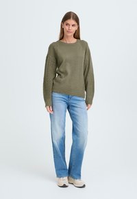 Olivgrön stickad tröja med texturerade ärmar, kombinerad med ljusblå raka jeans och beige sneakers. Minimalistisk design på en enfärgad bakgrund.