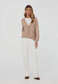 Donna con cardigan beige a bottoni, camicia bianca, pantaloni bianchi larghi e scarpe marroni, in piedi davanti a uno sfondo bianco semplice.