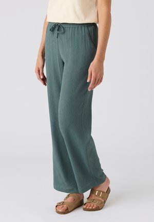 Persona con pantalones verdes holgados con una cintura elástica con cordón y sandalias beige con hebilla, de pie frente a un fondo liso.