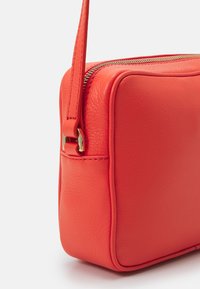 Rød skuldertaske i læder med tekstureret finish, rektangulær form, lynlås og en slank justerbar rem med gyldne metaldekorationer.