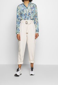 Floral shirt in blue, green, and beige; cream tapered pants with a belt and a front buckle; white sneakers with black accents.