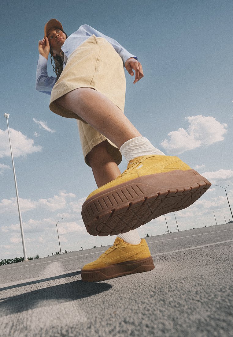 Zapatillas amarillas con una suela gruesa marrón, parte superior texturizada y calcetines blancos. Capturadas desde un ángulo bajo contra un fondo de cielo despejado.