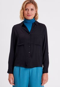 Chemise noire à manches longues en tissu léger, avec une coupe décontractée. Elle présente deux poches poitrine et des poignets à boutons.