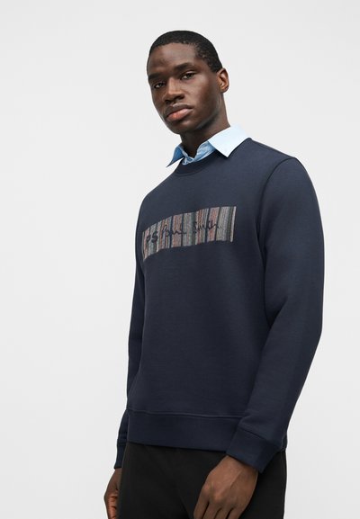 Άνδρας με μπλε σκούρο φούτερ με πολύχρωμο ορθογώνιο πάτς και ανοιχτό μπλε πουκάμισο με γιακά, μπροστά σε απλό λευκό φόντο.