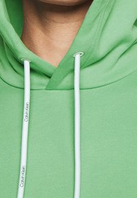 Felpa verde con coulisse bianca, nome del marchio Calvin Klein stampato sulla coulisse sinistra, inquadratura ravvicinata del collo e della parte superiore del torace.
