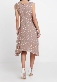 Braun gepunktetes Kleid aus leichtem Stoff, mit einem schichtweisen Rock, ärmellosem Design und einem Reißverschluss hinten. Weiße Absätze sichtbar.