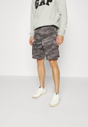 Homme portant un short cargo camouflage gris, des baskets blanches et un sweat-shirt gris clair avec le texte "GAP", debout devant un fond uni.