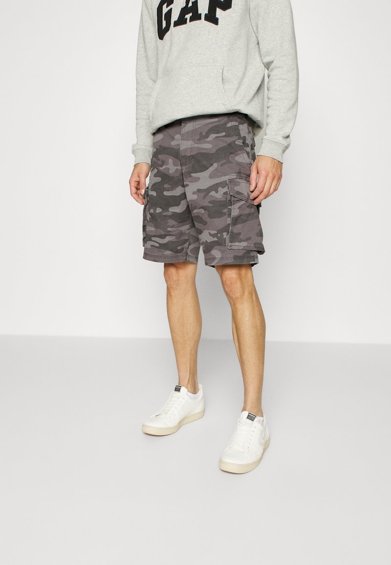 Homme portant un short cargo camouflage gris, des baskets blanches et un sweat-shirt gris clair avec le texte "GAP", debout devant un fond uni.