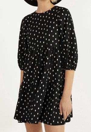 Woman wearing a black dress with white polka dots, three-quarter puff sleeves, and a tiered skirt, standing against a plain background.