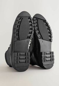 Schwarze Lederstiefel mit strukturierten Gummisohlen, die ein Profilmuster für guten Halt aufweisen. Das Design beinhaltet auffällige Nähte und minimalistische Akzente.