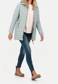 Mujer caminando con chaleco acolchado de color azul claro con capucha, camiseta de un tono rosa pálido, jeans ajustados azules y botas de tobillo marrones.