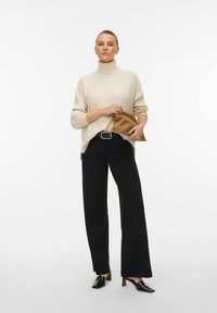 Bež turtleneck pulover, črne široke hlače, drži rjav kluček, črne mule s peto z teksturirano površino, minimalističen dizajn.