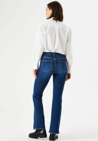 Blauwe denim flare jeans met een hoge taille, voorzien van achterzakken, een soepele textuur, en gedragen met een wit overhemd met knopen en zwarte laarzen.