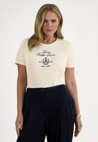 Εκρού βαμβακερό μπλουζάκι με κεντημένο ναυτικό λογότυπο και έμβλημα. Κοντά μανίκια και στρογγυλή λαιμόκοψη, συνδυασμένο με σκούρο παντελόνι.