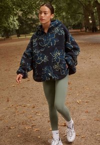 Niebieska bluza polarowa z kwiatowym wzorem, z kapturem, zamkiem z przodu i oversize’owymi rękawami; w połączeniu z oliwkowozielonymi legginsami i białymi sportowymi butami.