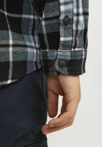 Camisa a cuadros en negro y azul con puños abotonados y una textura suave. Mano descansando sobre una tela oscura. Los botones son redondos y marrones.