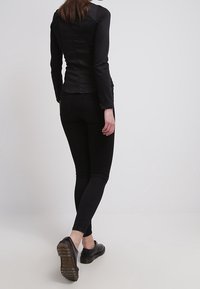 Top aderente nero a maniche lunghe con texture liscia, abbinato a jeans skinny neri e scarpe slip-on nere, visto da dietro.