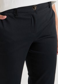 Pantalons noirs ajustés au toucher lisse, avec une poche avant, des plis cousus et un bouton doré décoratif à la taille.
