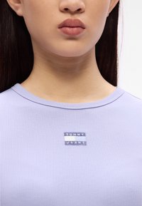Hellblau-lila gestreiftes T-Shirt mit rundem Halsausschnitt und einem kleinen aufgenähten rechteckigen Etikett, das "TOMMY JEANS" in Blau und Weiß zeigt.