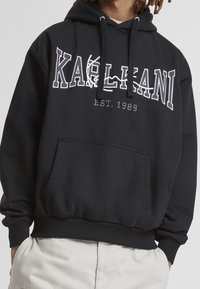 Svart huvtröja med framficka och vit text "KAIHLANI EST. 1989" tryckt på bröstet. Har ribbade mudd och nederkant.