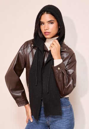 Hnedá kožená bunda so zipsom, krémový rolák a dlhý čierny pletený šál s kapucňou, s textúrovaným vzorom a strapcami na koncoch.